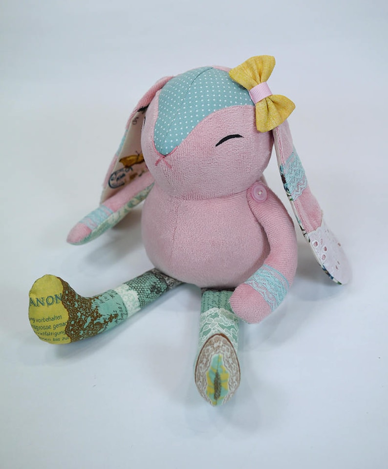 Puede incluir: Un conejito de peluche rosa hecho a mano con orejas de lunares azules y blancos, un lazo amarillo y patas de patchwork. El conejito est&aacute; sentado sobre un fondo blanco.