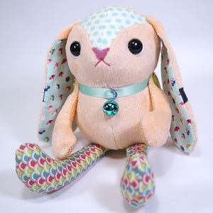 Puede incluir: Un juguete de peluche de conejo con un cuerpo rosa claro, orejas con lunares azules y patas con patrones coloridos. El conejo tiene un collar azul claro con una campanilla verde.