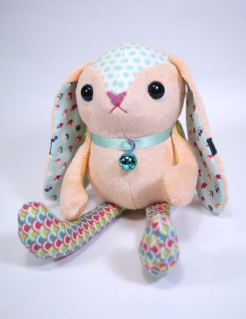 Puede incluir: Un juguete de peluche de conejo con un cuerpo rosa claro, orejas con lunares azules y patas con patrones coloridos. El conejo tiene un collar azul claro con una campanilla verde.