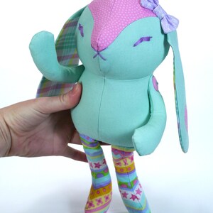Puede incluir: Un conejito de peluche azul claro con orejas de lunares rosas y un lazo morado. El conejito tiene orejas largas y ca&iacute;das y lleva pantalones a rayas de colores.