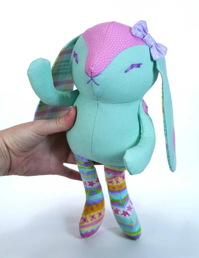 Puede incluir: Un conejito de peluche azul claro con orejas de lunares rosas y un lazo morado. El conejito tiene orejas largas y ca&iacute;das y lleva pantalones a rayas de colores.