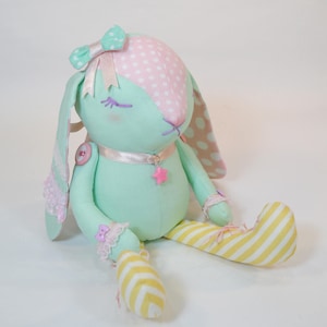 Puede incluir: Un conejito de peluche hecho a mano con un patr&oacute;n de lunares rosa y blanco en la cabeza y un cuerpo verde. El conejito tiene un lazo rosa, una cinta blanca alrededor del cuello y un amuleto de estrella rosa. El conejito est&aacute; sentado con las piernas cruzadas y lleva pantalones de rayas amarillas y blancas.