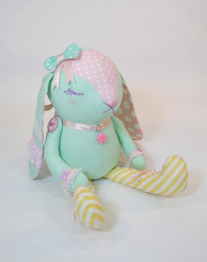 Puede incluir: Un conejito de peluche hecho a mano con un patr&oacute;n de lunares rosa y blanco en la cabeza y un cuerpo verde. El conejito tiene un lazo rosa, una cinta blanca alrededor del cuello y un amuleto de estrella rosa. El conejito est&aacute; sentado con las piernas cruzadas y lleva pantalones de rayas amarillas y blancas.