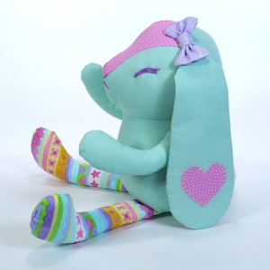Puede incluir: Un conejito de peluche azul claro con un coraz&oacute;n rosa en la espalda y un lazo morado en la cabeza. El conejito lleva polainas rayadas de colores.