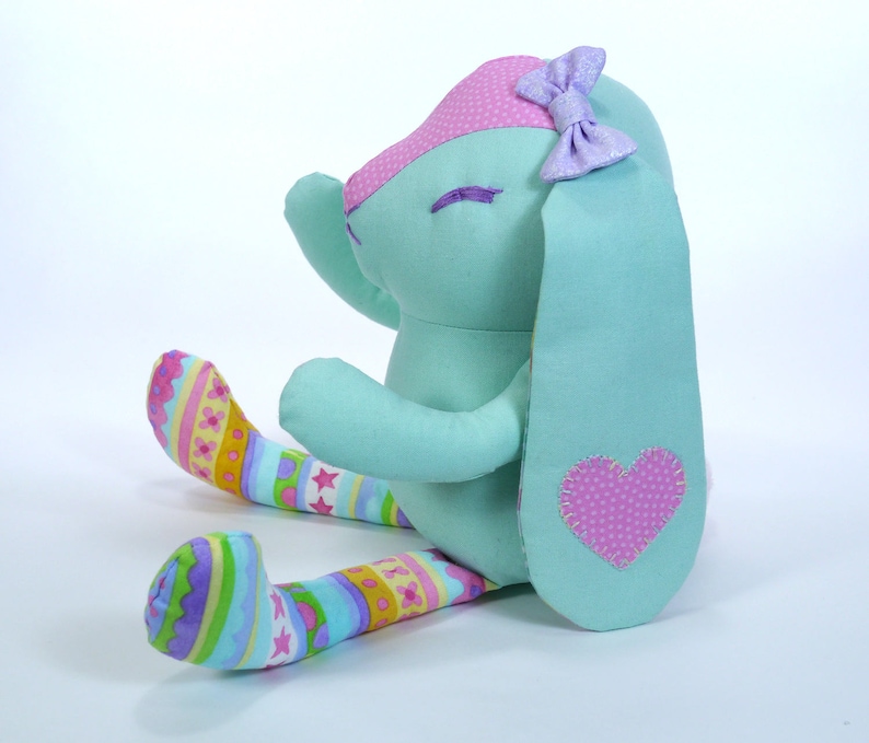 Puede incluir: Un conejito de peluche azul claro con un coraz&oacute;n rosa en la espalda y un lazo morado en la cabeza. El conejito lleva polainas rayadas de colores.
