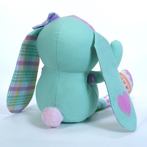 Puede incluir: Un suave conejito de peluche turquesa con un coraz&oacute;n rosa en una oreja, un lazo a cuadros y una cola de pomp&oacute;n rosa. El conejito sostiene un peque&ntilde;o huevo de Pascua colorido.