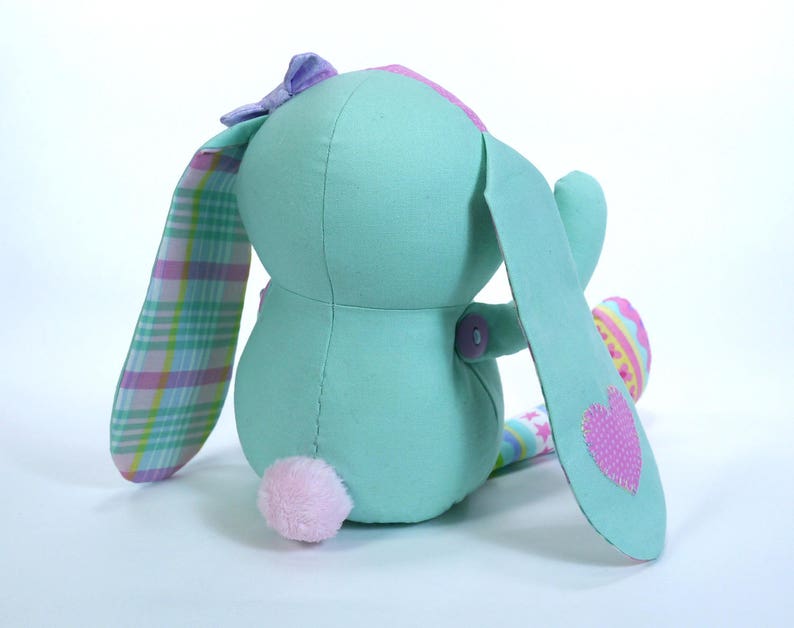 Puede incluir: Un suave conejito de peluche turquesa con un coraz&oacute;n rosa en una oreja, un lazo a cuadros y una cola de pomp&oacute;n rosa. El conejito sostiene un peque&ntilde;o huevo de Pascua colorido.