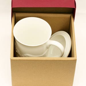 May include: A white ceramic mug with a handle and a matching saucer, packaged in a brown cardboard box with a burgundy lid.