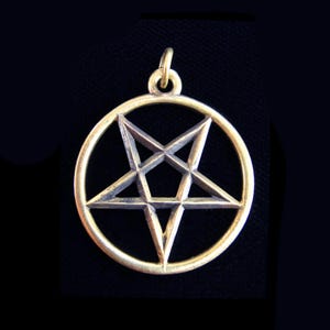May include: A gold-colored pendant featuring a pentagram within a circle. The five-pointed star is a prominent design element, with a loop at the top for a chain. The pendant is set against a dark background.