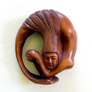 May include: A carved wooden sculpture of a figure in a circular pose, holding a small sphere. The figure's head is visible, with detailed facial features and a textured, flowing hair-like element. The wood has a warm, brown tone.