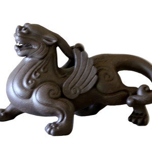 May include: A dark brown ceramic sculpture of a mythical creature with wings, a long tail, and intricate details. The creature is depicted in a crouching position with its head turned to the side.