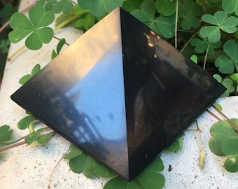 May include: A black obsidian pyramid with a reflective surface. The pyramid is sitting on a white surface with green leaves in the background.