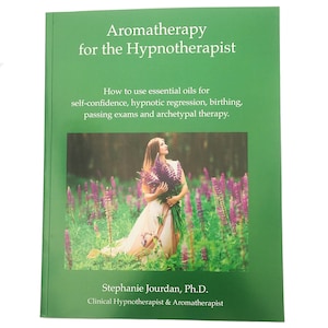 May include: A green book cover with the title "Aromatherapy for the Hypnotherapist" and a subtitle about using essential oils for self-confidence, hypnotic regression, birthing, passing exams, and archetypal therapy. The cover features a woman in a white dress holding a bouquet of purple flowers in a field.