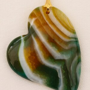 Puede incluir: Colgante en forma de coraz&oacute;n con una fianza dorada. El colgante presenta un patr&oacute;n en capas de colores verde, blanco y marr&oacute;n. La piedra tiene una superficie pulida y un aspecto natural y org&aacute;nico.