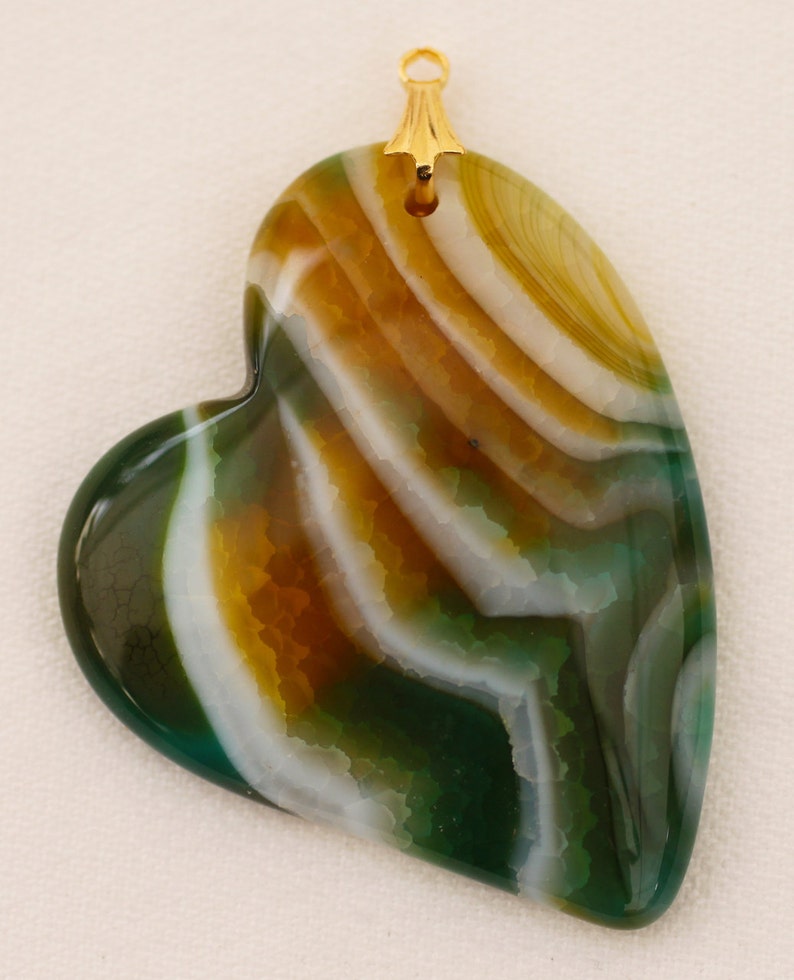 Puede incluir: Colgante en forma de coraz&oacute;n con una fianza dorada. El colgante presenta un patr&oacute;n en capas de colores verde, blanco y marr&oacute;n. La piedra tiene una superficie pulida y un aspecto natural y org&aacute;nico.