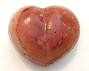 Puede incluir: Una piedra pulida en forma de corazón, en tonos naranja y marrón. La piedra tiene un acabado brillante y una superficie lisa. El corazón es un símbolo de amor y afecto, y esta piedra sería un gran regalo o artículo decorativo.