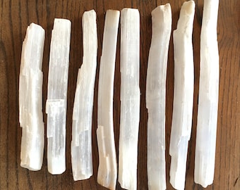 May include: Eight white selenite sticks, also known as selenite wands, are arranged in a row on a wooden surface. The sticks are smooth and have a natural, crystalline texture.