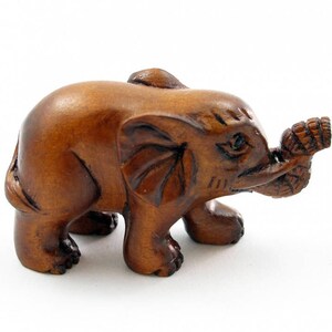 May include: A small, intricately carved wooden elephant figurine. The elephant is a warm brown color with detailed carvings on its ears and trunk. It is holding a bundle of leaves in its trunk. The figurine is standing on a white surface.