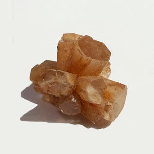 K&ouml;nnte beinhalten: Ein Cluster aus hellbraunen Aragonitkristallen. Die Kristalle sind durchscheinend, mit sichtbaren inneren Strukturen und einer warmen Farbpalette. Die Mineralformation ist vor einem wei&szlig;en Hintergrund platziert, was ihre nat&uuml;rliche Form hervorhebt.