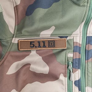 May include: Camouflage jacket in shades of green, brown, and blue. The jacket features a brown rectangular patch with the text "5.11" in dark blue. A green zipper and a small pocket are visible.