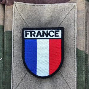 Peut inclure: Un écusson rectangulaire avec le mot "FRANCE" en lettres blanches au-dessus d'un design tricolore vertical bleu, blanc et rouge. L'écusson est bordé de noir et cousu sur un carré de tissu beige, sur un fond camouflage.