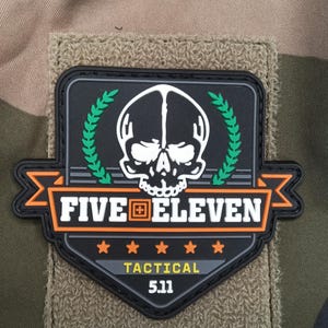 Peut inclure: Écusson noir et orange avec le texte "FIVE ELEVEN TACTICAL 5.11" et un motif de crâne. L'écusson est sur un fond en tissu beige et comporte une couronne de laurier verte.