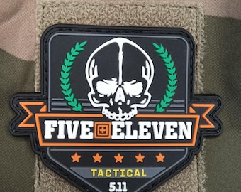May include: A black and orange patch with the text "FIVE ELEVEN TACTICAL 5.11" and a skull graphic. The patch is on a tan fabric background and features a green laurel wreath.