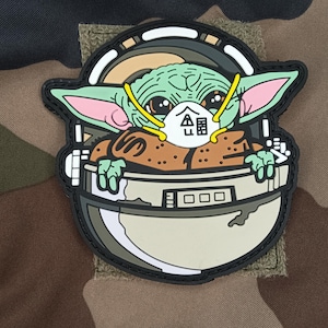 Op de afbeelding: Een patch met een groen, buitenaards personage met een gezichtsmasker, in een witte en grijze pod. Het personage heeft grote ogen en oren, met een bruinachtig gewaad. De patch staat op een camouflage achtergrond.