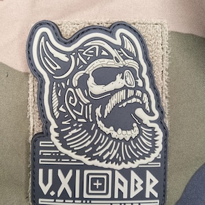 Puede incluir: Un parche gris y blanco con un cráneo vikingo estilizado con un casco con cuernos y una larga barba. El parche incluye el texto "V.XI.ABR" en letras blancas sobre un rectángulo gris. El parche está adherido a un cuadrado de tela beige.