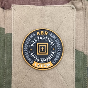 Peut inclure: Un écusson rond avec le texte "ABR TACTICAL" et "5.11 LATIN AMERICA LATAM" dans une police de style militaire. L'écusson est sur un carré de tissu beige, sur un fond de camouflage.