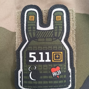 May include: A patch in the shape of a tactical vest, featuring the number "5.11" in white, a plus sign, and the word "PALEO" with a heart. The patch is primarily olive green with black and white accents, and is attached to a tan fabric.