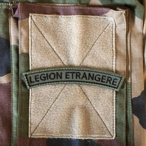 Puede incluir: Primer plano de un uniforme militar de camuflaje con un bolsillo y un parche rectangular. El parche dice "LEGION ETRANGERE" en letras negras sobre un fondo verde. El uniforme tiene un patrón de camuflaje marrón, verde y negro.