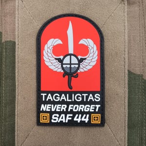 May include: A rectangular patch with a red background, featuring a white sword, wings, and a shield design. The text "TAGALIGTAS NEVER FORGET SAF 44" is displayed in white. The patch is set on a tan background with a camouflage pattern.