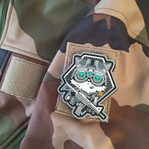 Può includere: Giacca mimetica con una toppa raffigurante un procione dei cartoni animati con un elmetto e un fucile. La toppa è grigia, bianca e nera con occhi turchesi. La giacca ha un motivo mimetico marrone, verde e beige.