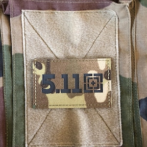 Pode incluir: Close-up de um tecido de camuflagem com um patch retangular. O patch apresenta o texto "5.11" em preto, com um design de mira. O tecido tem uma mistura de cores verdes, castanhas e bege.