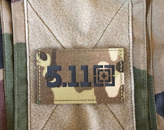 May include: Close-up of a camouflage fabric with a rectangular patch. The patch features the text "5.11" in black, with a crosshair design. The fabric has a mix of green, brown, and tan colors.