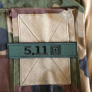 Peut inclure: Gros plan sur un tissu de camouflage avec une poche rectangulaire et un patch vert. Le patch affiche le texte "5.11" en noir, ainsi qu'un symbole plus. Le tissu présente des nuances de vert, de marron et de beige.