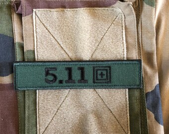 May include: Close-up of a camouflage pattern fabric with a rectangular pocket and a green patch. The patch features the text "5.11" in black, along with a plus sign symbol. The fabric has shades of green, brown, and tan.