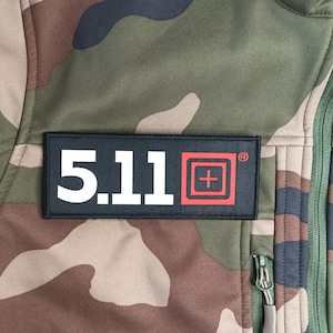 May include: Close-up of a camouflage jacket with a black rectangular patch. The patch features the text "5.11" in white, with a red square and cross logo. The jacket has a green zipper.