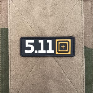 Peut inclure: Gros plan sur un patch avec le texte "5.11" en blanc et un carré jaune et noir avec un signe plus blanc. Le patch est sur un tissu beige, cousu sur un fond de camouflage.