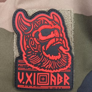 May include: A black and red patch featuring a stylized Viking head with a beard and horned helmet. The patch has the text "V.XI.ADR" in red. The patch is on a tan and green camouflage background.