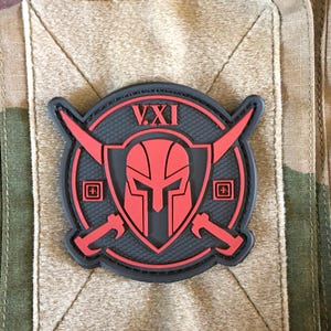 Op de afbeelding: Een zwart en rood embleem met een Spartaanse helm ontwerp, gekruiste bijlen en het Romeinse cijfer VXI. De patch is geplaatst tegen een camouflage achtergrond en heeft een getextureerd uiterlijk.