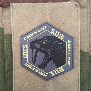 May include: A hexagonal patch with a snake design and the text "ALWAYS BE READY" and "5.11". The patch is dark blue with a black snake and gold lettering. It is attached to a tan fabric.