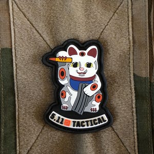 May include: A patch with a white cat design, incorporating red and orange details, holding a pencil and a stack of grey objects. The patch includes the text "5.11 TACTICAL" in white on a black border, set against a camouflage pattern.