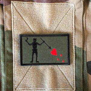 Op de afbeelding: Een rechthoekige patch met een zwart silhouet van een piraat met een speer, een rood hart en bloeddruppels. De patch staat op een groene achtergrond met een zwarte rand, tegen een vierkant van bruin en camouflage stof.