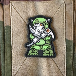May include: A green and black patch depicting a cartoon rhino in military gear, holding a knife and a tool. The rhino wears a green helmet and uniform. The patch is on a tan square against a camouflage background.