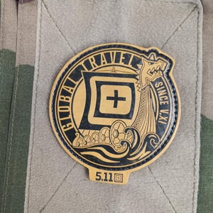 Peut inclure: Écusson beige et noir avec les mots "GLOBAL TRAVEL" et "SINCE V.XI" encerclant un symbole de croix. L'écusson présente également un motif de navire viking et le chiffre "5.11". L'écusson est cousu sur un tissu beige.