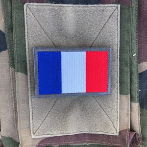 Peut inclure: Un écusson rectangulaire avec le drapeau français bleu, blanc et rouge est fixé sur un carré de tissu beige. L'écusson est cousu sur un fond à motif de camouflage.