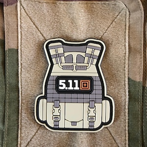 May include: A patch featuring a stylized tactical vest design. The patch is primarily gray, tan, and black, with the text "5.11" and a small orange square. The patch is attached to a camouflage fabric.