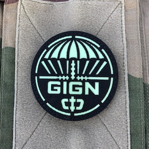 Peut inclure: Un écusson rond noir avec un motif de parachute et le texte "GIGN" en blanc. L'écusson est posé sur un fond de camouflage beige et vert. Le parachute est vert clair.